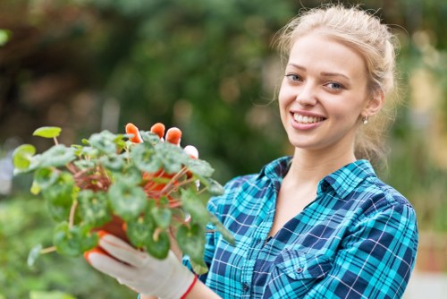 A young woman with blonde hair tied back, wearing a blue checked shirt and gardening gloves, is tending to a potted plant with green and red leaves in an outdoor garden. The garden features a well-maintained lawn area with dense green grass, bordered by hedges and shrubs. In the background, there are taller trees with leafy canopies, providing partial shade, and a paved patio section with wooden decking. The scene appears to be during daylight with natural sunlight illuminating the garden space, indicating favourable weather conditions. This outdoor area demonstrates a thoughtfully landscaped yard suitable for gardening activities, with clear attention to plant care and garden maintenance, aligning with professional gardening and landscaping services provided by Landscapers Tooting in the local area near SW17 postcode.