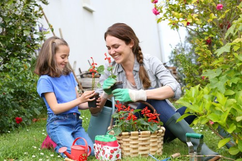 A woman and a young girl are gardening outdoors in a well-maintained backyard in Tooting, with lush green grass, flowering plants, and shrubs surrounding them. The woman is smiling as she waters a potted red flowering plant, holding a small watering can, while the girl, wearing a blue t-shirt and jeans, holds the same potted plant and appears to be assisting. They are seated on the grass among gardening tools, including a basket with additional pots, a gardening glove on the woman's hand, and a small red watering can. The garden features a variety of green foliage and flowering shrubs in the background, with natural daylight illuminating the scene, emphasizing a peaceful outdoor environment suitable for planting, lawn care, and garden maintenance tasks, often managed by local gardening services in Tooting.