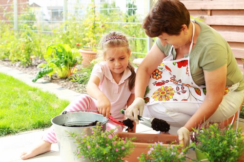 A woman and a young girl are working together in a well-maintained garden in Tooting, engaged in planting or gardening activities. The woman, wearing a light green top and a floral apron, is kneeling on the patio and handling dark soil or compost in a rectangular planter box. The girl, dressed in a light pink top and striped trousers, is sitting beside her with a small gardening trowel, actively assisting with planting. In the foreground, there are vibrant pink flowering plants in pots, enhancing the outdoor space’s visual appeal. The background features a lush, green garden with a variety of plants, shrubs, and a vegetable patch enclosed with a wire fence, indicating a productive and well-organized garden environment. The patio surface appears to be wooden decking, with a garden bucket nearby. The natural lighting and slightly overcast sky create a gentle, inviting atmosphere suitable for outdoor gardening activities, reflecting the professional and caring approach of local gardening services such as Landscapers Tooting.