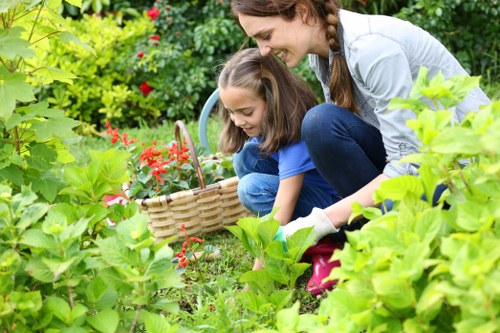 A woman and a young girl are engaged in gardening activities in a lush, well-maintained backyard. The woman is crouching down beside the girl, both focused on planting or tending to plants in a flower bed. The garden features a dense arrangement of green foliage, with a variety of shrubs and leafy plants, some with vibrant red flowers. Behind them, there are taller bushes and possibly a hedge, providing privacy and structure to the outdoor space. The lawn area in the foreground has well-kept grass with a lush, healthy appearance, and the flower bed is bordered with natural soil and a few scattered pebbles. Sunlight illuminates the scene, suggesting a clear day suitable for outdoor gardening. The garden environment exudes a tidy, natural look typical of residential gardens in Tooting, South London, reflecting local gardening styles. The natural setting is ideal for general gardening and outdoor lawn care services offered by Landscapers Tooting, emphasizing their expertise in maintaining vibrant, attractive outdoor spaces.
