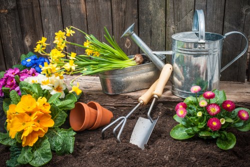A close-up view of a prepared garden bed in a backyard, featuring dark, rich soil with freshly turned areas. Surrounding the bed are colorful flowering plants including pink and yellow primroses, along with a pot of yellow primulas. A weathered wooden fence encloses the garden, providing privacy and a natural backdrop. In the background, various garden tools such as a watering can, a hand rake, and a trowel rest on the soil, indicating active planting and maintenance efforts. A small, empty terracotta pot is also visible atop the soil near the flowers. The overall scene depicts a well-maintained outdoor space suitable for gardening services, with natural lighting suggesting a partly cloudy day. The garden environment showcases a combination of flowering plants, soil preparation, and garden tools, reflecting typical landscaping and gardening activities relevant to Tooting and the local area.
