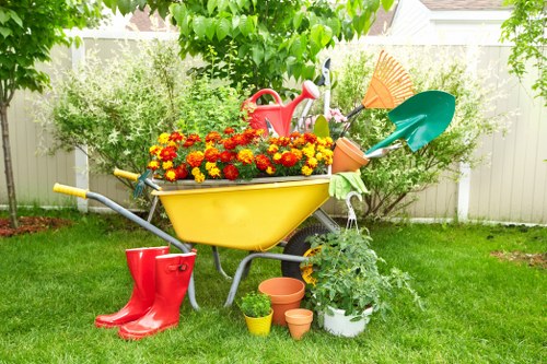 A colourful garden scene featuring a yellow wheelbarrow filled with vibrant red and yellow flowers situated on a well-maintained, lush green lawn. Surrounding the wheelbarrow are various gardening tools and accessories, including a red watering can placed in the wheelbarrow, a pair of red rubber boots on the grass nearby, and multiple plant pots in different sizes and terracotta shades positioned on the ground. Behind the wheelbarrow is a hedge with dense, leafy foliage, and two small trees or shrubs help frame the background, providing defined garden borders. The scene appears to be set during a bright, sunny day with natural light illuminating the garden, creating a fresh and inviting outdoor space typical of residential landscaping in Tooting. This image exemplifies gardening setup and outdoor maintenance, aligning with typical garden care services offered in the local area by Landscapers Tooting.