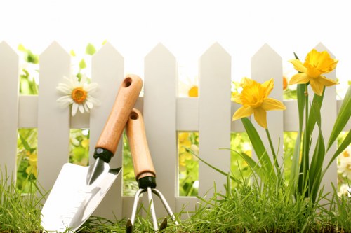 A close-up view of a small garden area in Tooting, featuring a white picket fence in the background, with a variety of garden tools including a trowel, secateurs, and a small garden fork placed on the green grass foreground. To the right, there are blooming yellow daffodils and white daisies adding natural colour to the scene. Behind the fence, lush green foliage indicates a well-maintained outdoor space, with sunlight illuminating the flowers and tools, suggesting a bright spring or summer day. The garden layout shows a neat, organized small yard suitable for outdoor gardening activities typical of residential homes in Tooting, supporting professional lawn maintenance and gardening services provided by Landscapers Tooting.