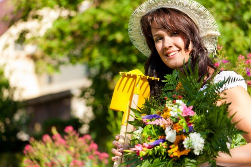 A woman standing outdoors in a landscaped garden area during daytime, holding a small yellow garden rake in one hand and a large bouquet of various colorful flowers and green foliage in the other. She is wearing a lightweight, wide-brimmed straw hat with decorative edging, and a casual white top. The garden features a well-maintained lawn with lush, densely grassed areas in the foreground, bordered by flower beds with a variety of blooms, and a backdrop of mature trees and shrubbery with green foliage. The scene is brightly lit, indicating clear weather, with sunlight casting natural light across the space. The setting suggests an outdoor gardening or planting project, highlighting the lush green tones of the lawn and the vibrant colors of the flowers, consistent with professional gardening services in Tooting, South London, provided by Landscapers Tooting.