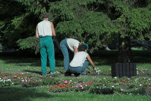 A gardener and two children working together in a lush, green garden with mature trees providing shade. The garden features a well-maintained lawn with a variety of flowering plants arranged in beds along the front, showcasing pink, red, and white blossoms. The children are kneeling on the grass, tending to the plants, while the adult stands nearby, supervising or assisting. In the background, large leafy trees form a canopy over the outdoor space, and a black outdoor chair is visible on the lawn. The setting appears to be a peaceful residential garden in Tooting with natural light, ideal for gardening and outdoor maintenance activities, typical of professional landscaping services by Landscapers Tooting.