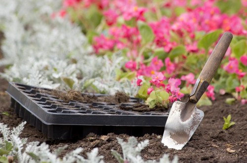 A gardening scene in a landscaped outdoor space in Tooting, showcasing a close-up view of freshly turned soil with a partially filled plastic seed tray on the ground. The soil appears dark and rich, prepared for planting, with some residual dirt indicating recent work. Bright pink and white flowers in full bloom are visible in the background, along with green foliage and small shrubbery, suggesting a well-maintained garden area. A garden trowel with a wooden handle is partially inserted into the soil, hinting at ongoing planting or soil preparation activities typical of professional gardening services. The environment is illuminated by natural daylight, with weather conditions indicating a clear or partly cloudy sky, adding to the vibrant natural tones of the garden scene. This detailed image highlights the tools, plant beds, and soil management involved in outdoor gardening and landscaping aimed at enhancing residential or commercial outdoor spaces in Tooting, as part of comprehensive lawn care and garden maintenance performed by Landscapers Tooting.