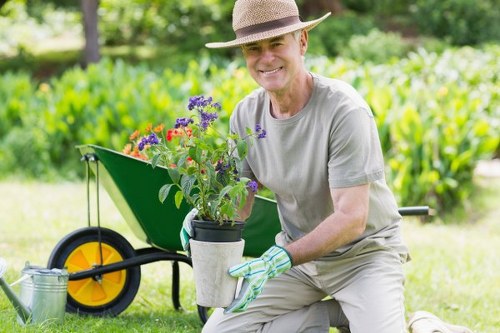 A mature man wearing a wide-brimmed straw hat, light grey t-shirt, and gardening gloves is kneeling on a well-maintained lawn in a backyard garden in Tooting. He is holding a small plant with purple and red flowers in a grey protective container, showing a gentle smile. Behind him, there is a green garden wheelbarrow with bright yellow wheels, positioned on a paved area adjacent to a lush, green hedge and a grassy lawn. In the background, neatly trimmed shrubs and trees provide a natural setting, with the bright sunlight illuminating the scene and casting soft shadows. The scene depicts an outdoor space suitable for gardening and landscaping services, with a focus on sustainable and eco-friendly gardening practices, aligning with the 'Recycling and Sustainability' theme at landscaperstooting.co.uk.