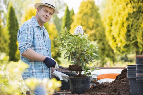 In the image, a man wearing a light-colored straw hat, a blue checked shirt, and gardening gloves is working in an outdoor garden space. He is holding a small plant in a black plastic pot, preparing to plant it into the soil. The garden features a wooden bench or table with several empty black plant pots stacked nearby, alongside some gardening tools and a small pile of soil. In the background, there is lush greenery with tall trees and well-maintained hedges, indicating a landscaped yard with a mix of lawn, shrubbery, and flower beds. The scene is set on a bright, sunny day with natural light illuminating the garden, suggesting active gardening or landscaping work by a professional service such as Landscapers Tooting, who offer garden maintenance and planting services in the local area of Tooting, London. The environment appears clean and organized, highlighting the qualities of a well-kept outdoor space suitable for gardening and landscaping improvements.