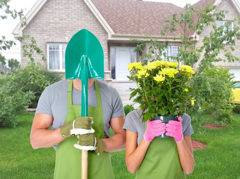 A person and a child are standing in a neatly maintained front garden of a suburban house, both wearing green aprons and gardening gloves; the adult is holding a garden trowel with a blue plastic protective cover, while the child is holding a large pink pot filled with vibrant yellow flowers, obscuring their faces. The garden features a lush, well-kept green lawn, bordered by small shrubs and young trees, with a pathway leading to the house's entrance, which has light-colored brick walls and white-framed windows. The background includes a two-storey house with a sloped roof and a garden bed with soil and planted greenery. The scene suggests outdoor gardening activity on a bright, clear day, emphasizing lawn care, planting, and garden maintenance services typical of a professional landscaper in Tooting or nearby London areas, reflecting a well-designed, family-friendly garden environment.