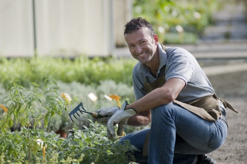 A man with short dark hair and a broad smile is kneeling in a well-maintained garden, surrounded by greenery and flowering plants. He is wearing a light grey shirt, blue jeans, and a brown gardening apron, and is holding a small hand rake in his right hand, positioned over a bed of lush, green foliage with small yellow and white flowers. The garden features a tidy lawn area, bordered by a narrow pathway made of compacted soil or gravel, and is enclosed by a wooden fence in the background. Behind him, there is a gentle slope with additional plantings and trees, indicating a thoughtfully landscaped outdoor space. The lighting suggests a bright, partly sunny day, with natural daylight illuminating the scene. This outdoor garden setting exemplifies typical features of residential gardening and landscaping in Tooting, South London, aligning with professional gardening services offered by Landscapers Tooting.